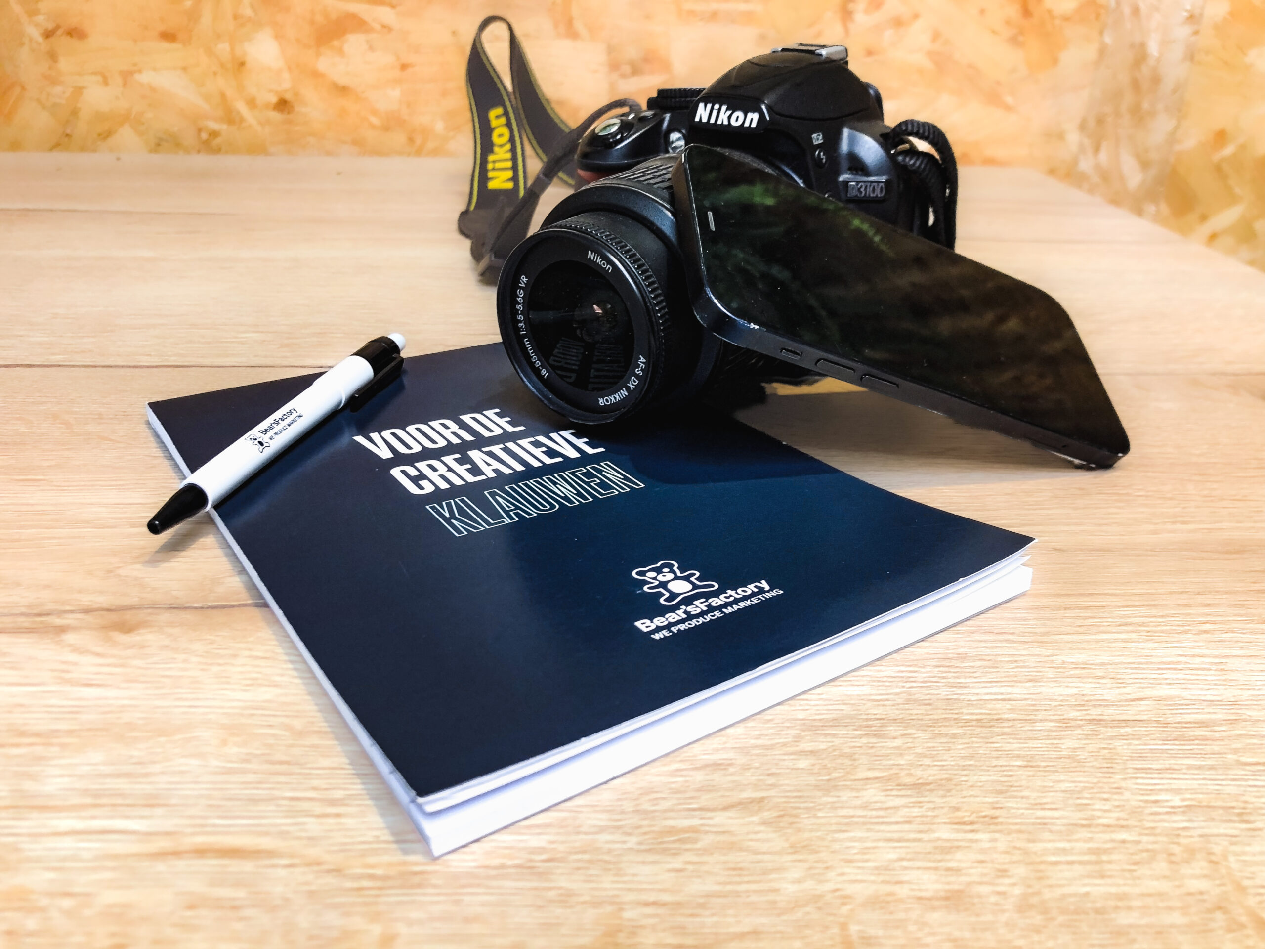 Compositie van Bear's Factory notitieblok en pen, Nikon camera en telefoon.